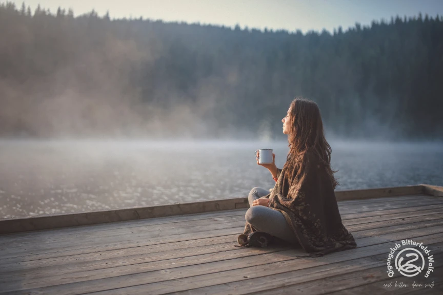 Frau mit einer Tasse auf einem Holzsteg am nebligen See, mit dem Logo des Qigongclubs Bitterfeld-Wolfen